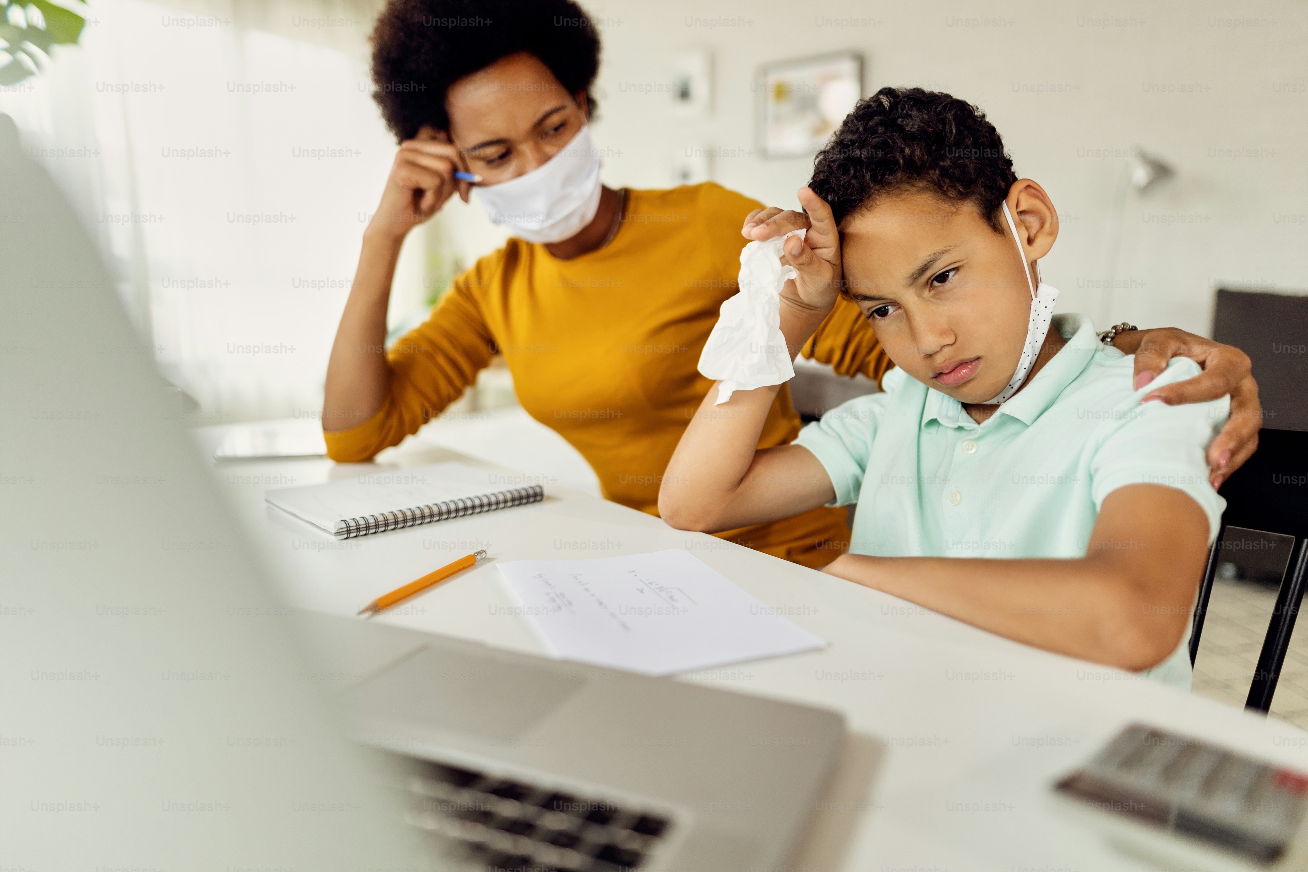Black boy feeling displeased while e-learning over laptop with his mother during COVID-19 pandemic.