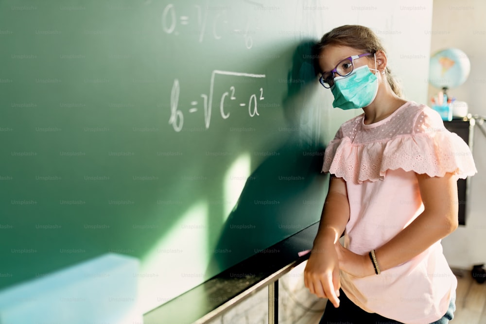 Schoolgirl with protective face mask feeling sad because she can't solve math assignment on blackboard.