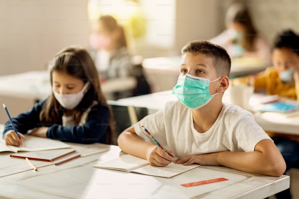 Schoolboy wearing protective face mask and raising arm to ask a question while learning with classmates in the classroom.