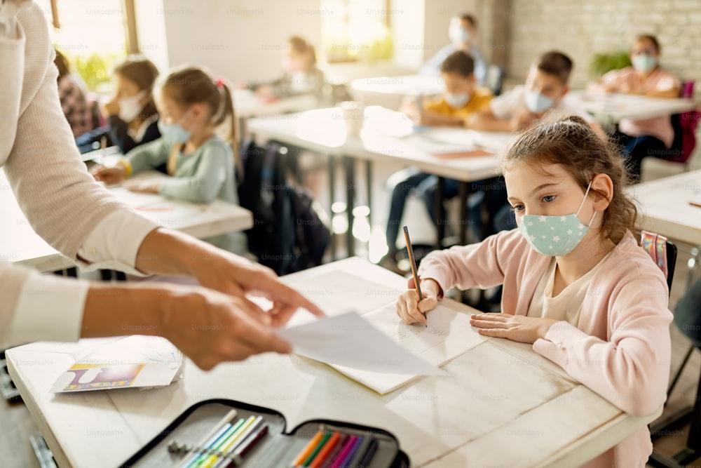 Kleines Mädchen trägt eine schützende Gesichtsmaske und schreibt, während ihr Lehrer ihr bei der Aufgabe im Klassenzimmer hilft.