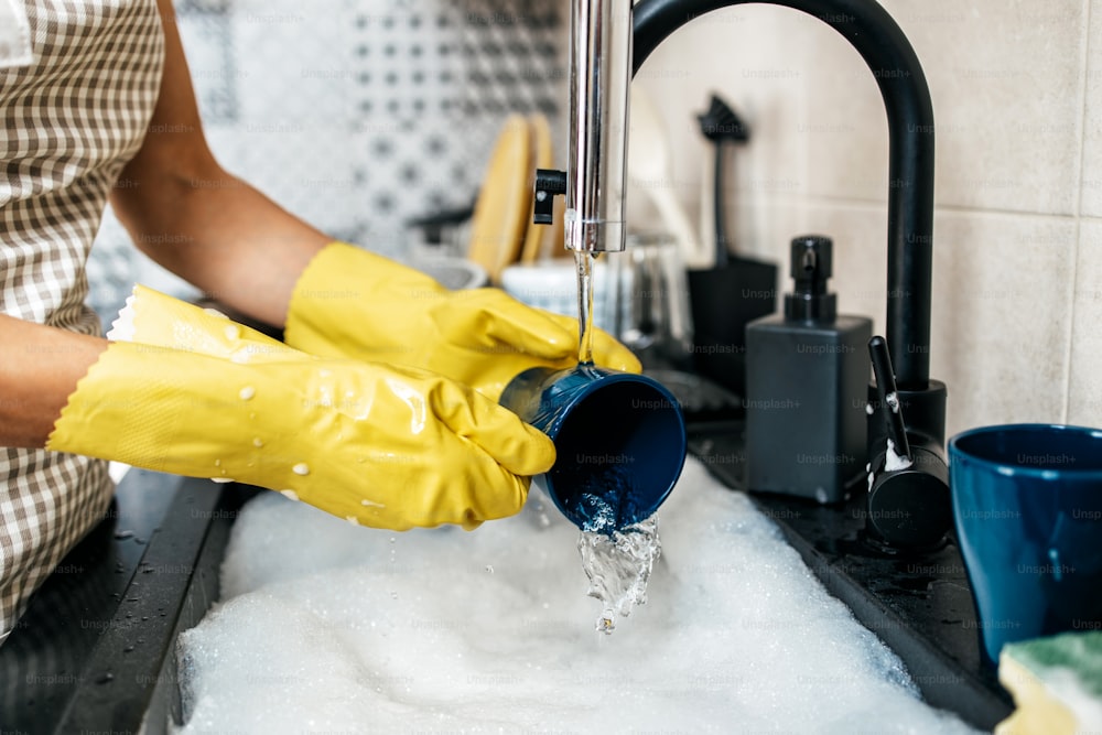 Young adult woman with yellow protective gloves washing her dishes on kitchen sink. Household and home hygiene routine.