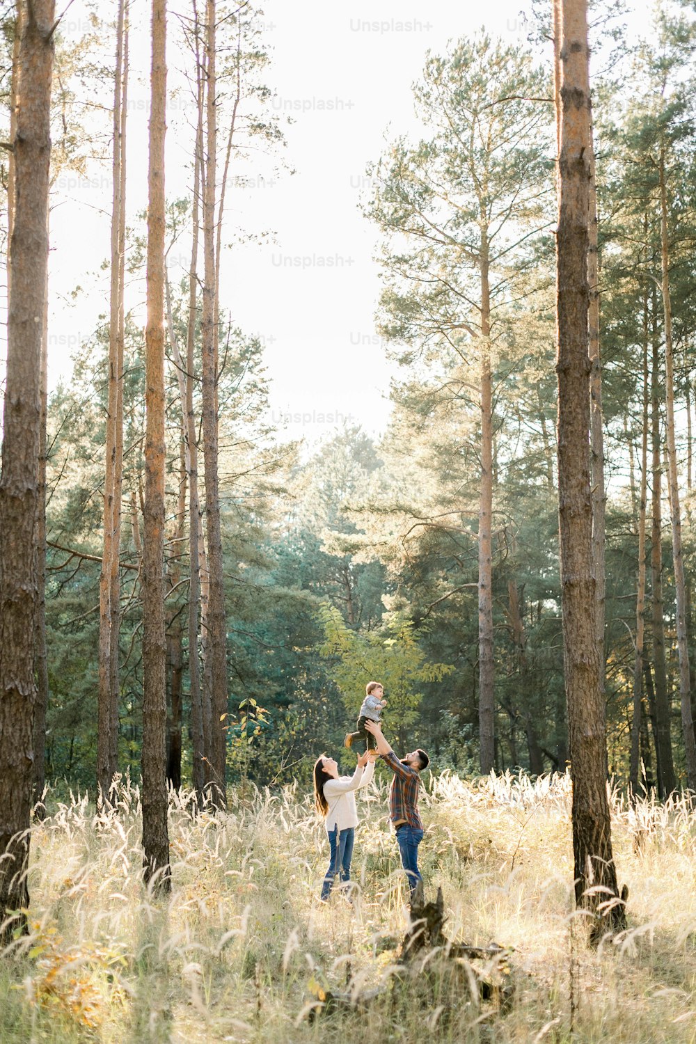 Outdoor portrait of happy young parents, having fun and lifting up their little cute baby son, during walk in autumn forest at sunny day.