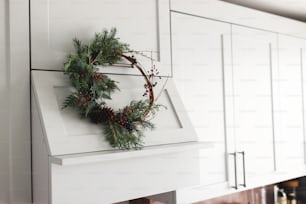 Rustic christmas wreath hanging on modern kitchen hood, festive holiday decoration. Creative modern christmas wreath hanging on stylish kitchen cabinets. Merry Christmas and Happy Holidays