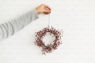 Christmas wreath in florist hand on white background, holiday advent. Female hand holding simple christmas wreath with red berries, rustic creative decor