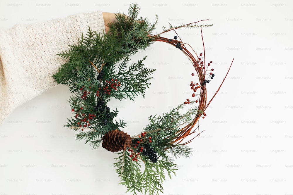 Rustic christmas wreath in female hands on white background. Woman in stylish knitted sweater holding modern christmas wreath with fir branches, berries and cone, holiday advent.