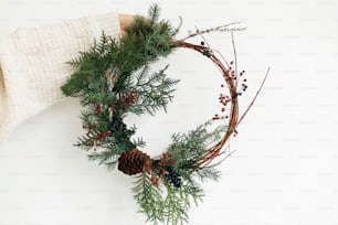 Rustic christmas wreath in female hands on white background. Woman in stylish knitted sweater holding modern christmas wreath with fir branches, berries and cone, holiday advent.