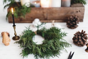 Corona navideña rústica con velas, piñas, hilo y adornos sobre mesa de madera blanca. Hacer una corona navideña simple y elegante con ramas de cedro, adviento del taller de vacaciones