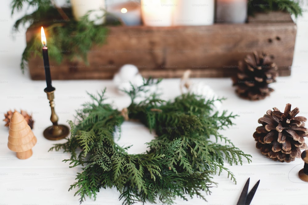 Rustikaler Weihnachtskranz mit Kerzen, Tannenzapfen, Faden und Ornamenten auf weißem Holztisch. Einfacher stilvoller Weihnachtskranz mit Zedernzweigen, Ferienwerkstatt Advent
