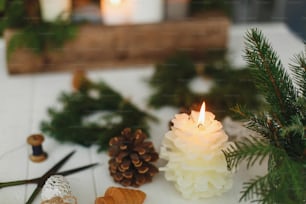 Burning candle in pine cone shape on rustic background with christmas wreath, pine cones and ornaments on wooden table in evening. Holiday advent, cozy atmosphere. Copy space