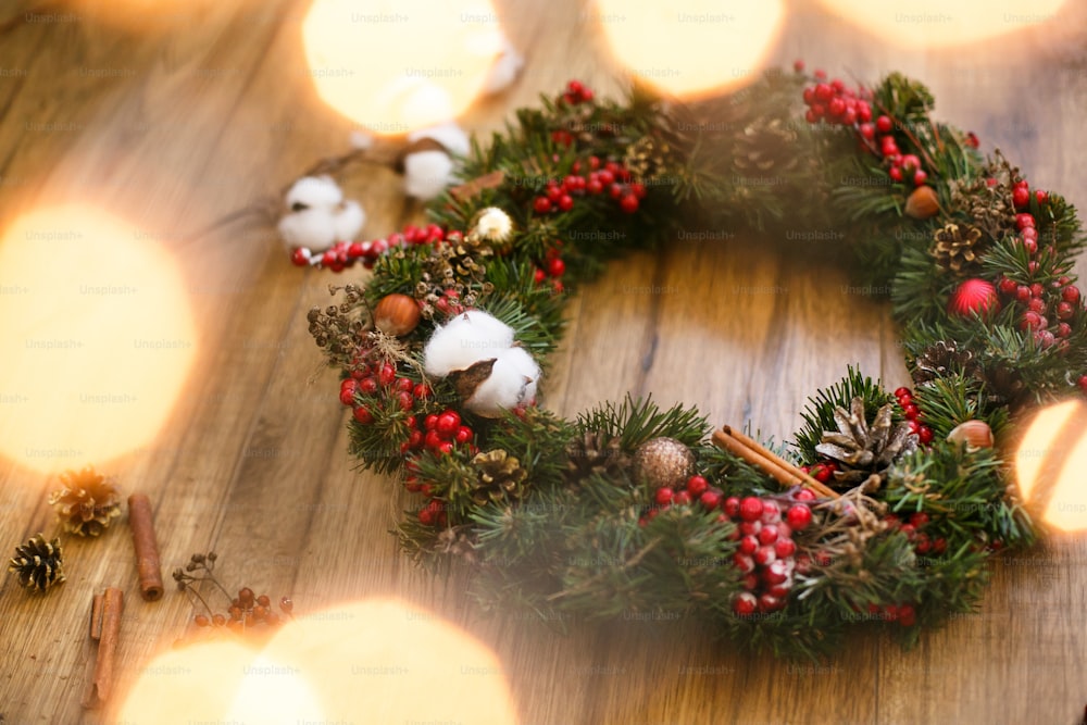 Weihnachtskranz auf rustikalem Holzhintergrund in Lichtern. Traditioneller Weihnachtskranz mit roten Beeren und Ornamenten, Tannenzapfen, Baumwolle und goldenen Lichtern auf Holz, Urlaubsdekor