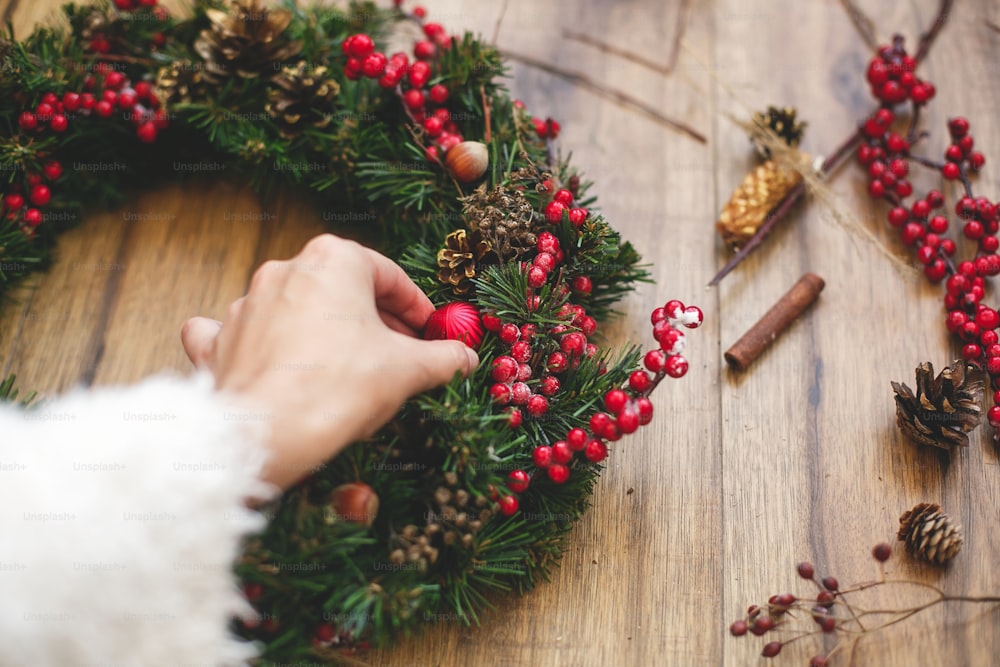 Person, die traditionellen Weihnachtskranz auf rustikalem Holzhintergrund herstellt. Florist schmückt Weihnachtskranz mit Ornamenten, Baumwolle, roten Beeren und Tannenzapfen. Ferienwerkstatt