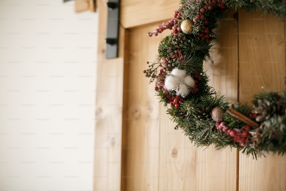 家の素朴な木製のドアにぶら下がっているクリスマスリース。木製の背景に赤い果実と装飾品、松ぼっくりとシナモン、休日の装飾と伝統的なクリスマスリース。テキスト用のスペース