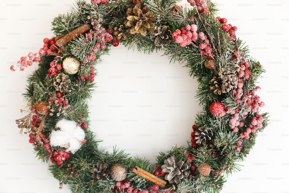 Weihnachtskranz hängt an weißer Wand im Haus. Traditioneller Weihnachtskranz mit roten Beeren und Ornamenten, Tannenzapfen und Zimt isoliert auf weißem Urlaubsdekor. Platz für Text