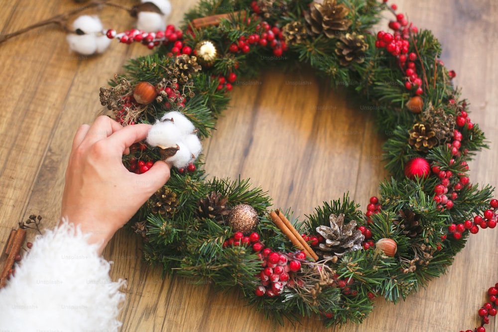 Person, die traditionellen Weihnachtskranz auf rustikalem Holzhintergrund herstellt. Florist schmückt Weihnachtskranz mit Baumwolle, roten Beeren, Ornamenten und Tannenzapfen. Ferienwerkstatt
