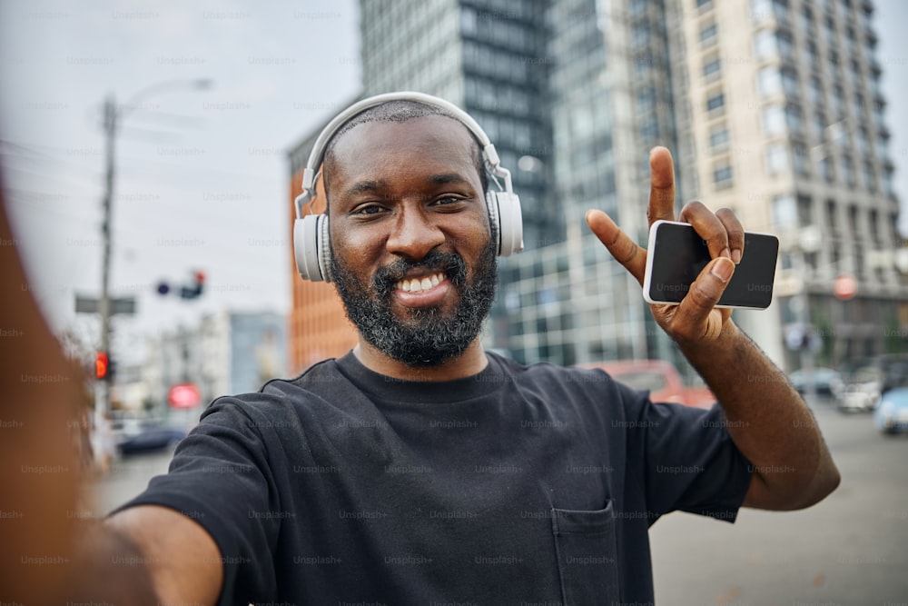 Cheerful bearded young man wearing headphones holding a modern smartphone and showing peace gesture with a happy smile