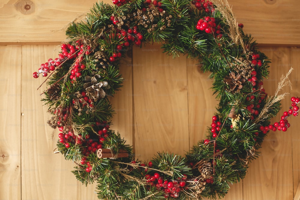Corona de Navidad que cuelga en la puerta de madera rústica de la casa. Corona navideña tradicional con frutos rojos y adornos, piñas y canela sobre fondo de madera, decoración navideña.