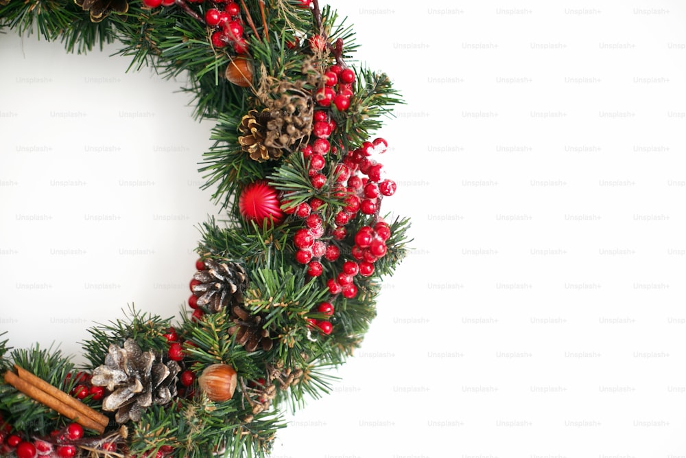 Coroa de Natal pendurada na parede branca em casa. Coroa de Natal tradicional com bagas vermelhas e enfeites, pinhas e canela isolada no branco, decoração natalina. Espaço para texto