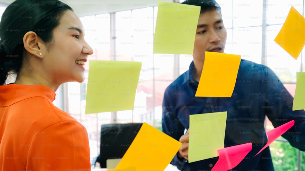 Business people work on project planning board in office and having conversation with coworker friend to analyze project development . They use sticky notes posted on glass wall to make it organized .