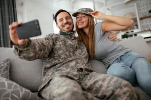 Jeune soldat prenant un selfie avec sa femme à la maison. Couple aimant assis sur le canapé dans le salon.