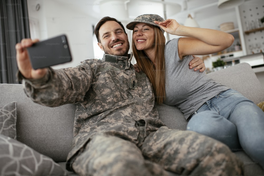 Young soldier taking selfie with his wife at home. Loving couple sitting on sofa in living room.