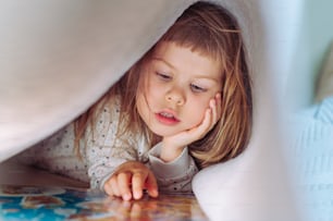 Beautiful little girl reading book lying on the bed under the blanket in the bedroom.