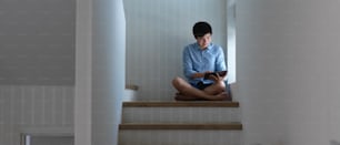 A man crossed legs sitting on the floor at stair in his home and using digital tablet