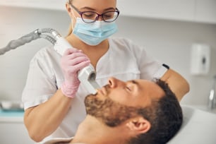 Bearded young man lying on daybed while female cosmetologist removing unwanted hair from his cheek with laser device