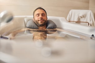 Caballero guapo con los ojos cerrados relajándose en la bañera y sonriendo mientras se somete a un procedimiento de hidroterapia