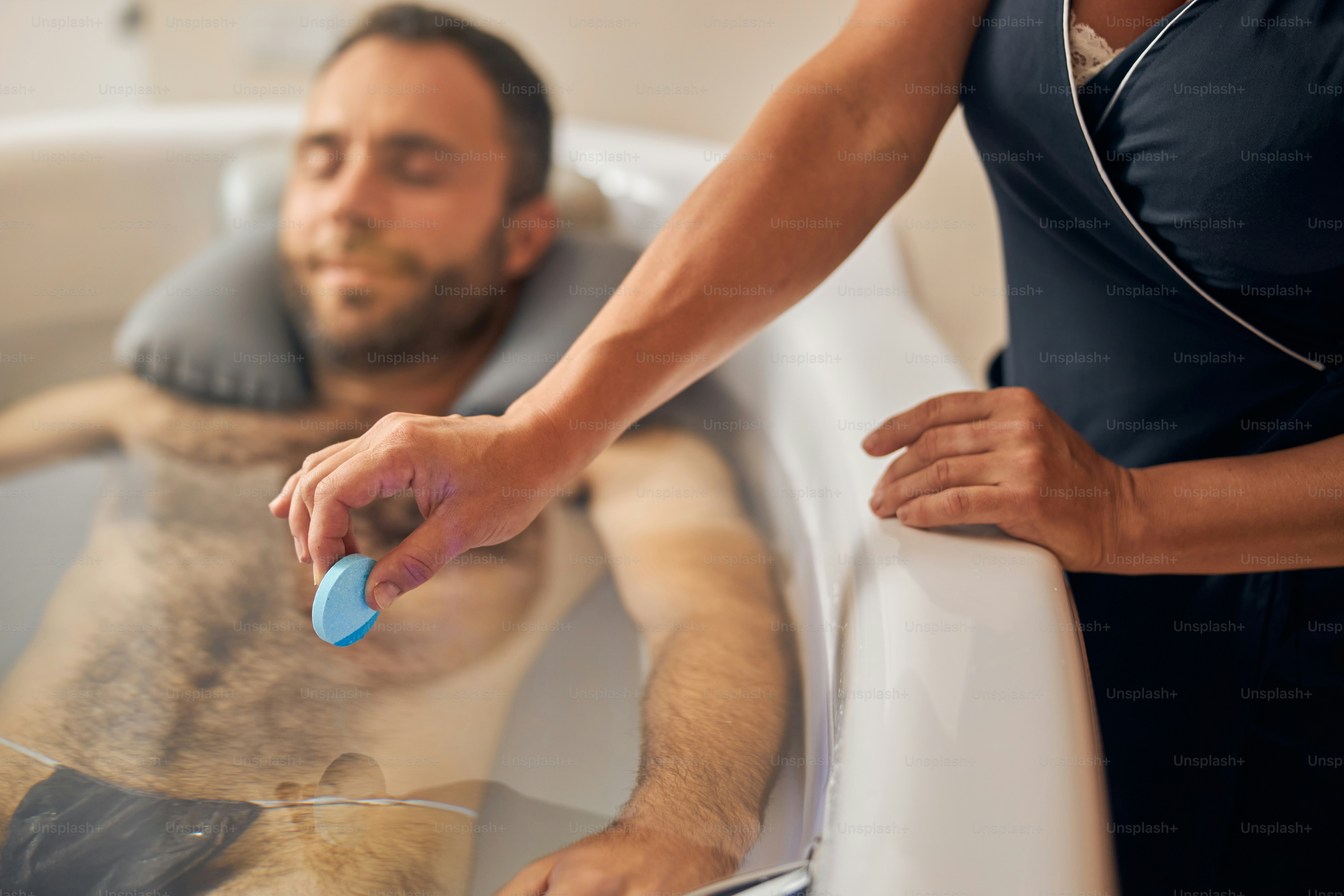 Handsome gentleman lying in bathtub while beautician putting relaxing bath tablet into water
