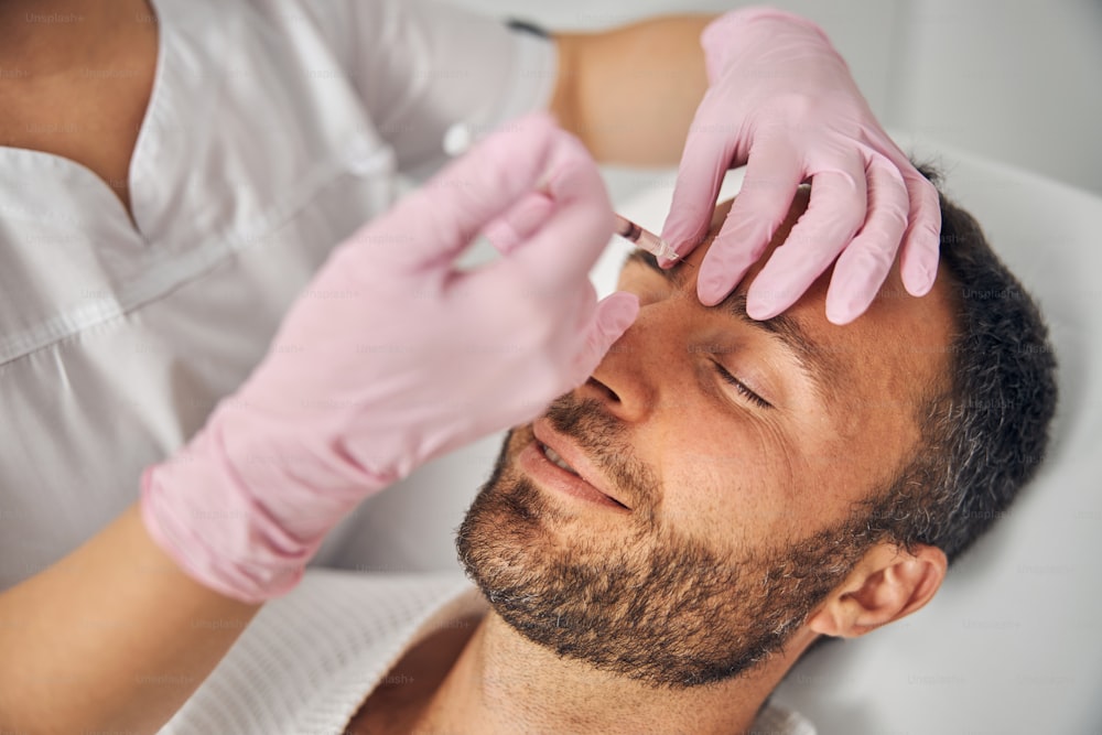 Nahaufnahme einer Ärztin der Kosmetikerin in sterilen Handschuhen, die eine Injektion in die männliche Stirn vornehmen