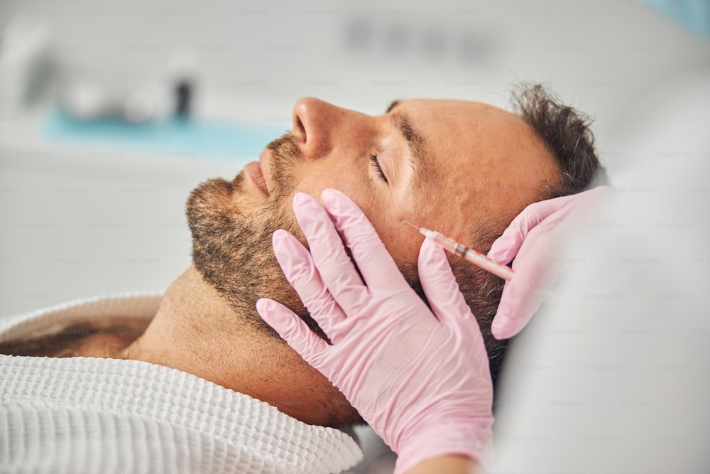 Gros plan sur les mains d’une cosmétologue féminine portant des gants stériles faisant une injection dans le visage d’un homme