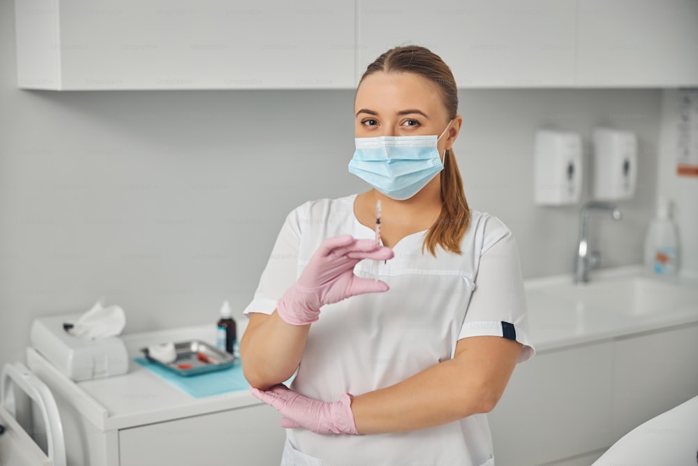 Charming young woman beautician with syringe in her hand wearing protective face mask while standing in cosmetology cabinet
