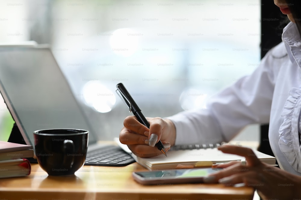 Closeup mulher mão trabalhando em um smartphone e escrevendo em um bloco de notas com uma caneta no escritório.