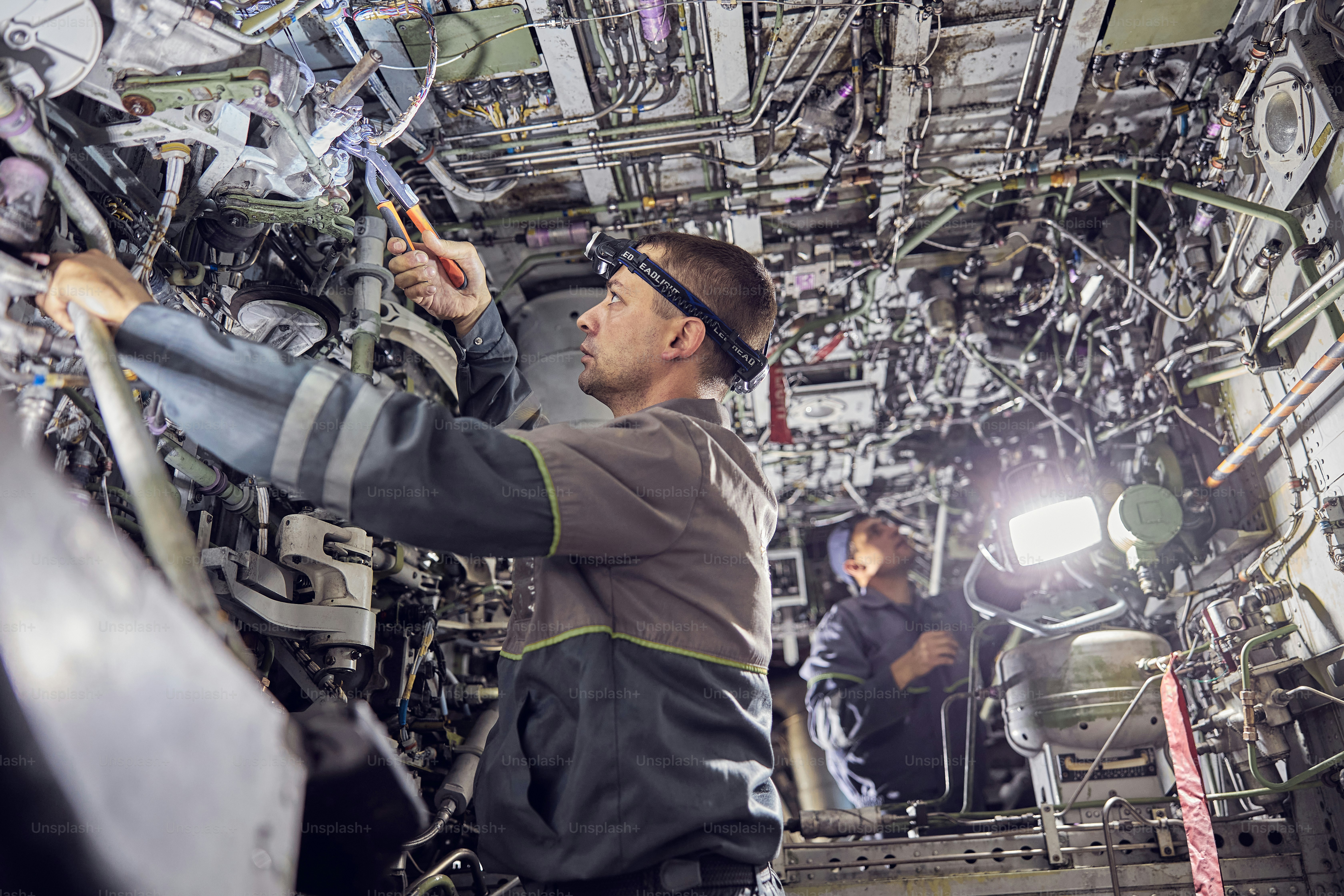 Retrato de vista lateral de dos mecánicos profesionales que visten uniforme general utilizando herramientas de aviación profesionales mientras están de pie dentro de la aeronave