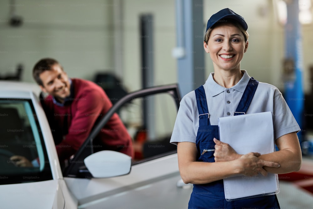 Porträt einer glücklichen Automechanikerin, die in einer Werkstatt steht und in die Kamera schaut. Im Hintergrund steht ein Kunde.
