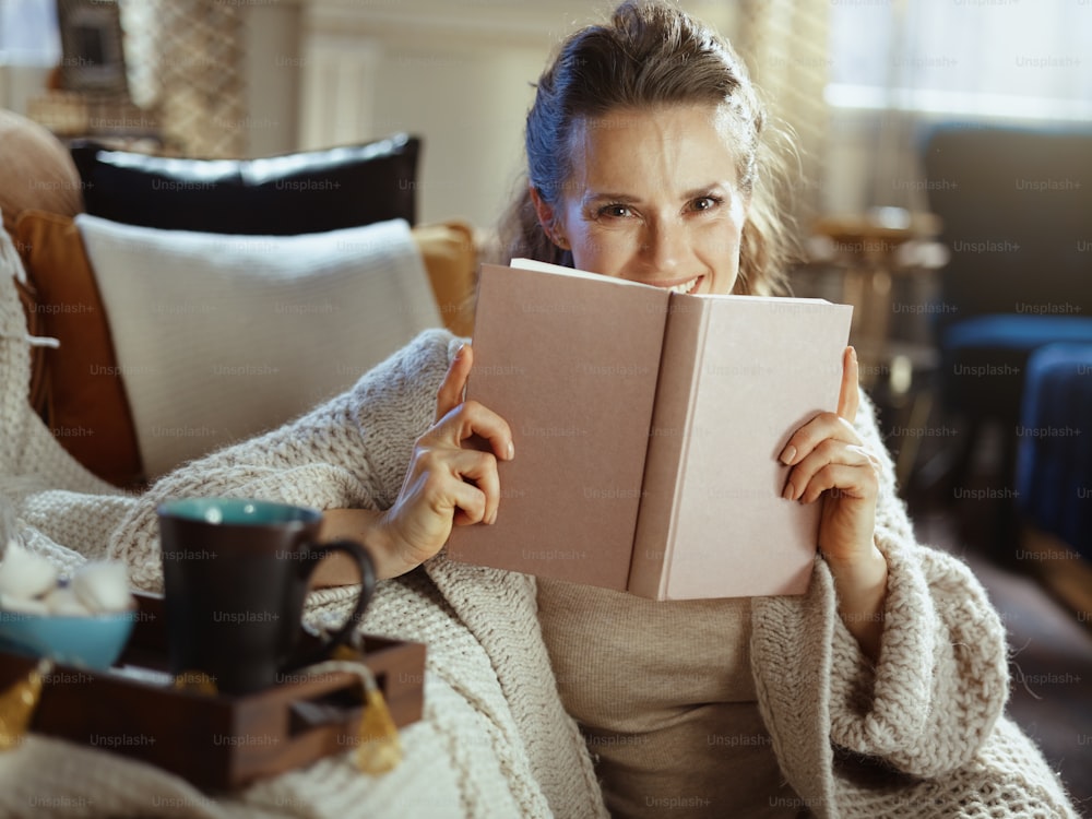 happy young female in knitted cosy cardigan with tray and cup reading book in the modern living room in sunny winter day.