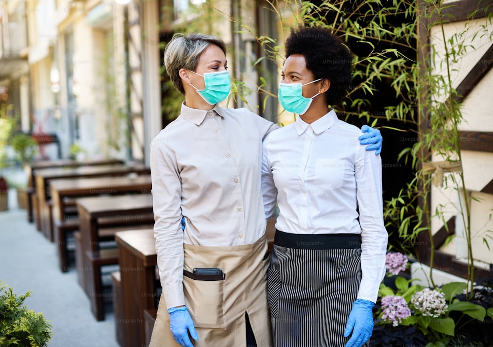 Happy waitresses standing embraced and talking while wearing protective face masks at sidewalk cafe.