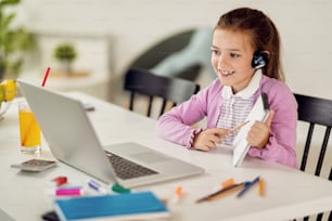Niña feliz usando la computadora y hablando con su maestra a través de una videollamada mientras aprende en casa.
