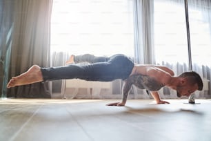 Side view portrait of sporty strong male in grey pants prepping for competition while doing hard elements of pole dance in class