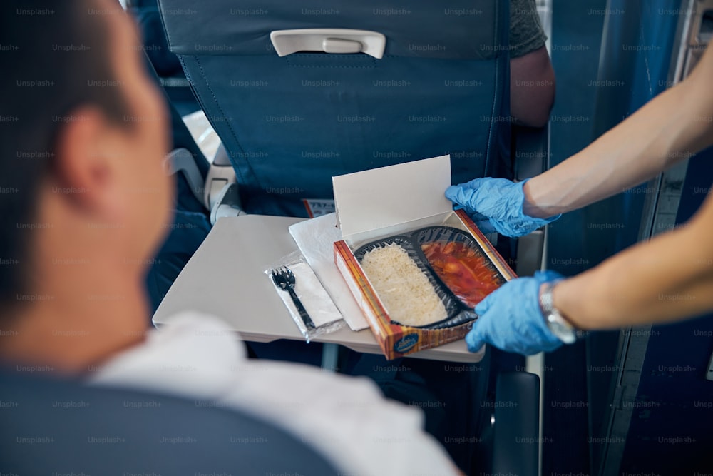 Close up portrait of air stewardess serving box of food for male passenger in protective gloves to prevent covid virus affection in airplane cabin