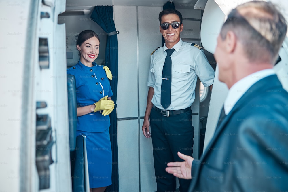 Cheerful pilot and stewardess are greeting elegant man on stairway while he is boarding before flight