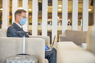 Hombre maduro con gafas y traje está usando un cuaderno mientras está sentado con máscara de seguridad y guantes cerca del equipaje en el aeropuerto