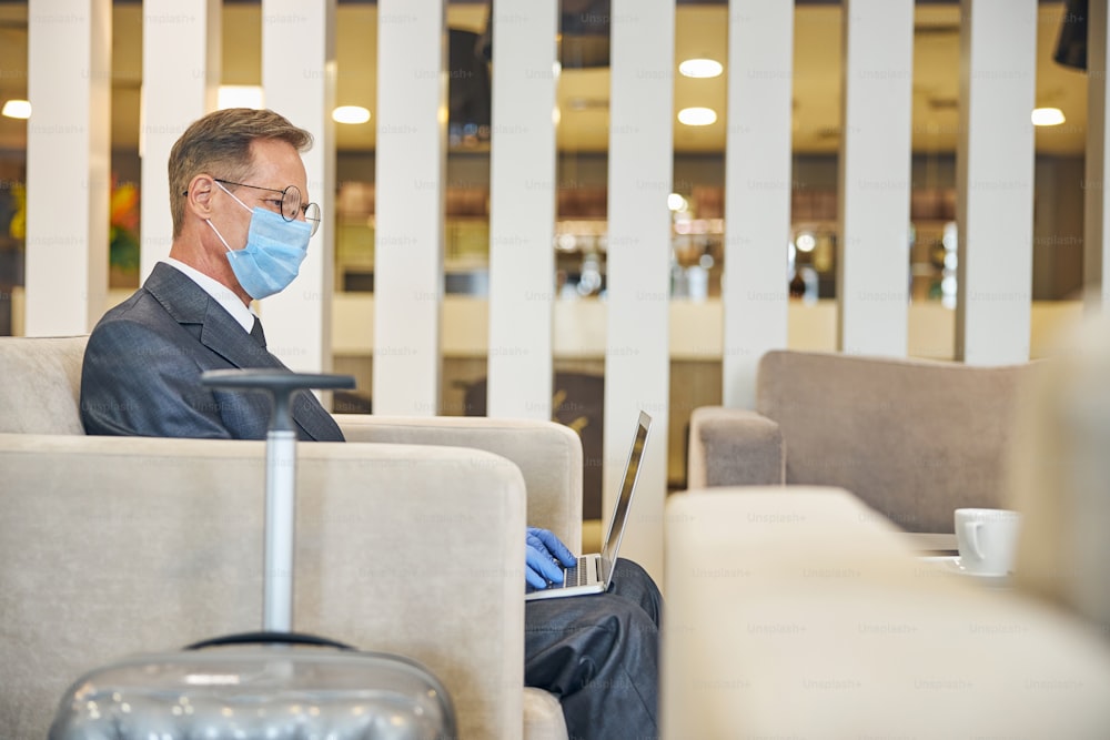 Mature male in glasses and suit is using notebook while sitting in safety mask and gloves near baggage in airport