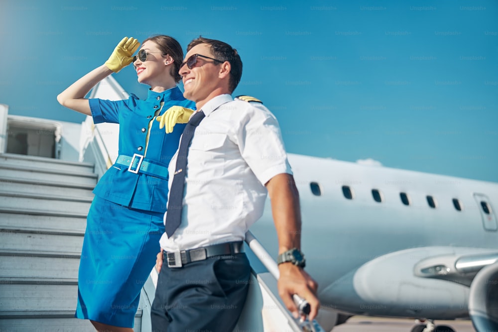 Low angle of happy young pilot and pretty stewardess standing together on stairway after landing