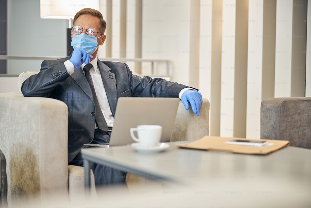 Elegener reifer Mann in Dlasses mit Schutzhandschuhen und Maske, während er vor dem Flug Notebook benutzt und Kaffee trinkt