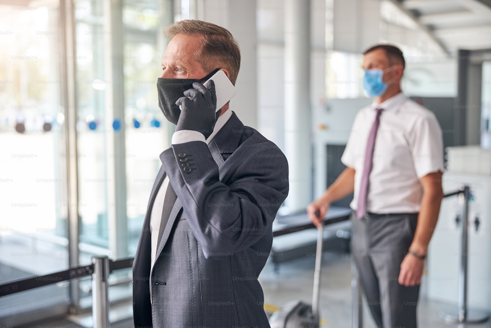 Geschäftsmann mit Schutzhandschuhen und Maske telefoniert, während Assistent Gepäck am Flughafen trägt