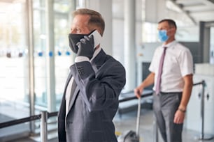 Businessman with protective gloves and mask is talking on cell phone while assistant carrying luggage in airport