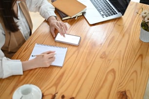 Cropped shot of desingner female writing her project on notebook while using smartphone with white screen on wooden desk.