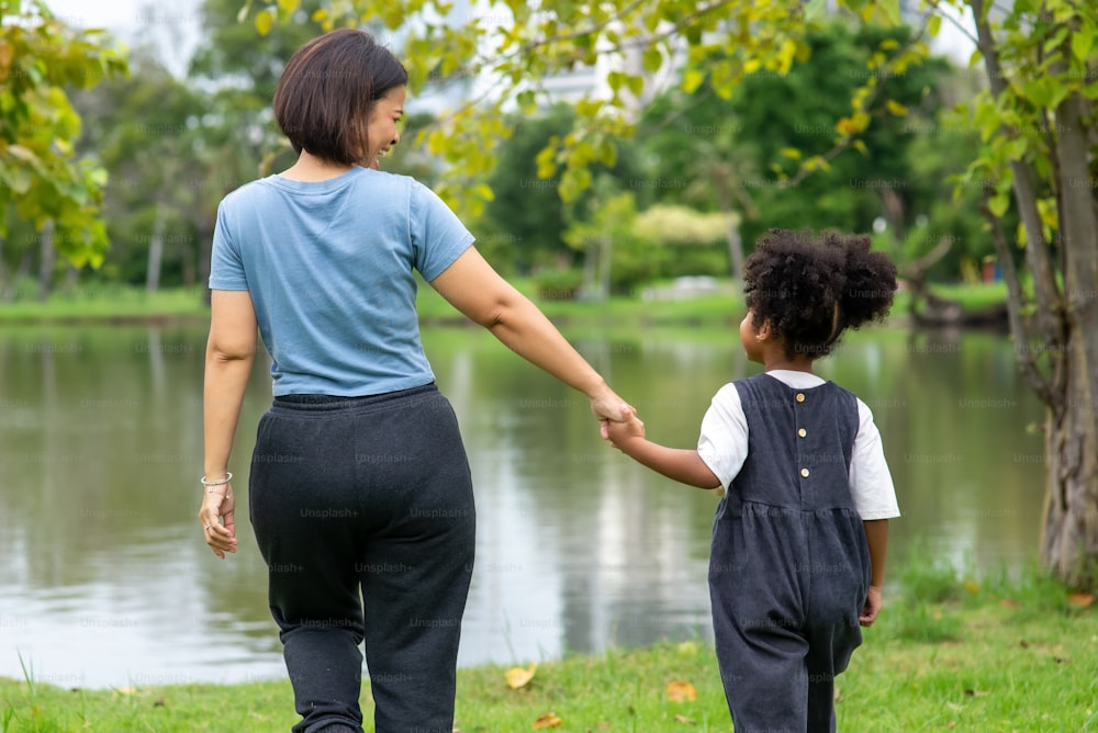 Rear view of Happy mixed race mother with little daughter holding hands and walking together in the park.