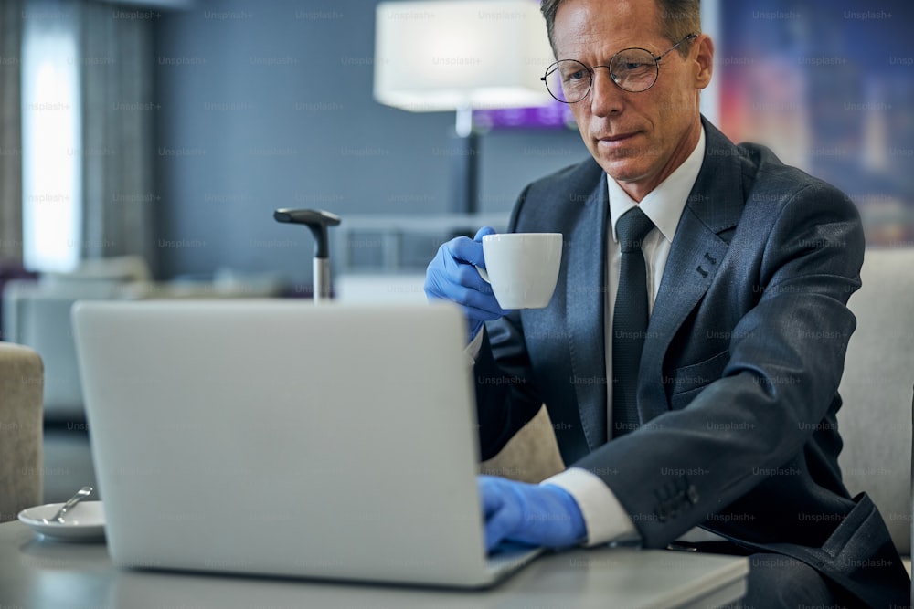Ernsthafter Mann in Anzug und Brille, der während der Pandemie vor dem Flug Kaffee trinkt und Notebook benutzt